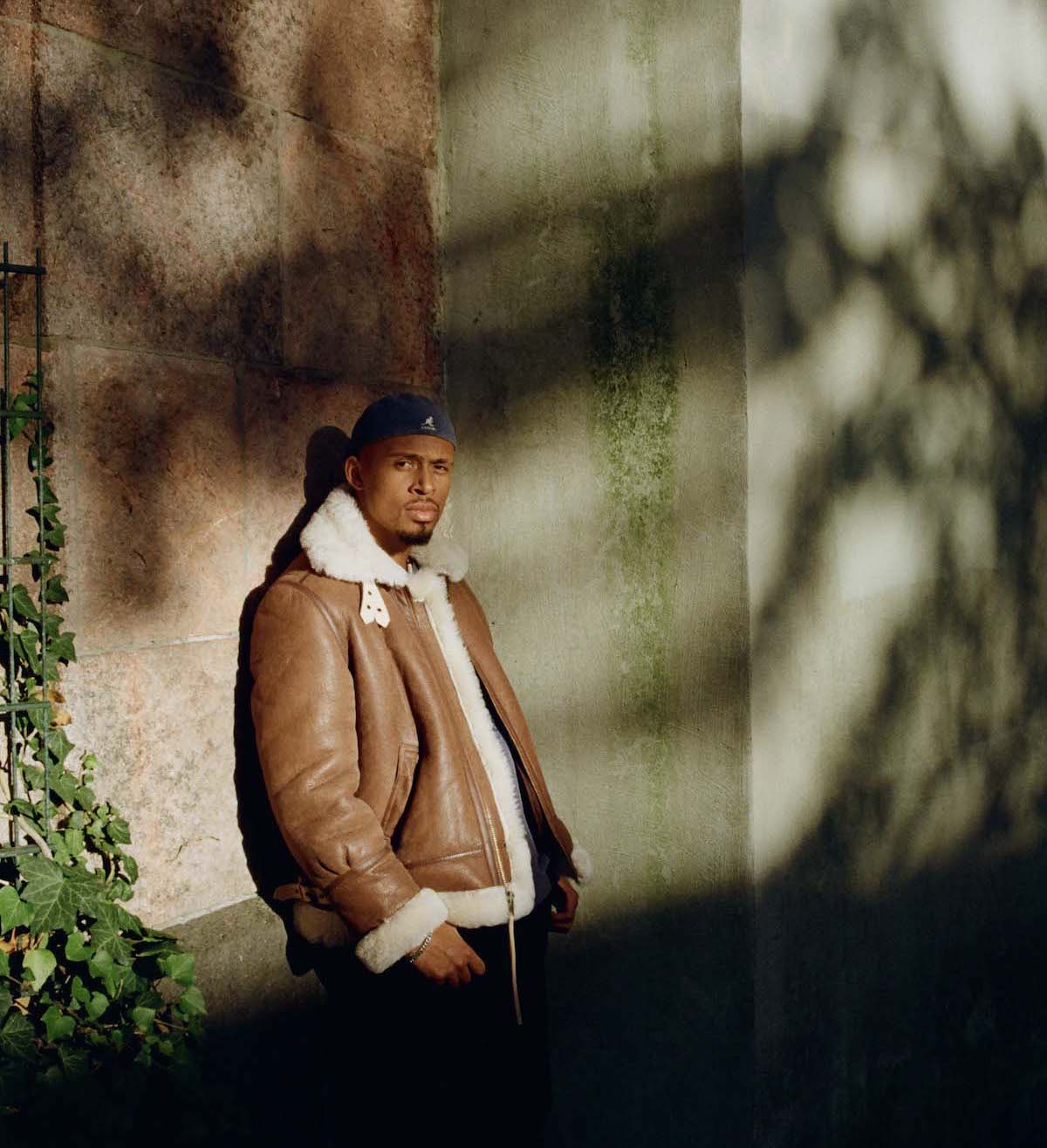Black, young man with moustache and goatee, wears a blue-grey bonnet, a brown, thick leather jacket with fur collar and fur trim on the sleeves and waistband, his thumbs are in his trouser pockets. Ansu looks into the camera. He is leaning against a high stone wall with ivy growing on it. The sun is shining, shadows of a tree fall on the scene.