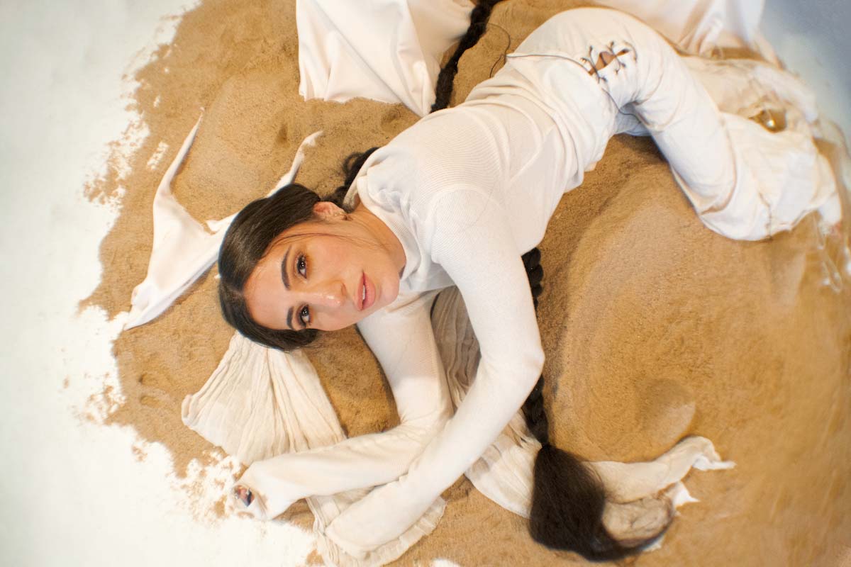 Young woman with long, braided pigtail lies on her side on a pile of sand heaped on top of a white fabric that can still be seen at the edge. Evîn is photographed from above, looking into the camera. She is wearing a white long-sleeved shirt, the sleeves of which she has also pulled over her hands, and white cloth trousers.