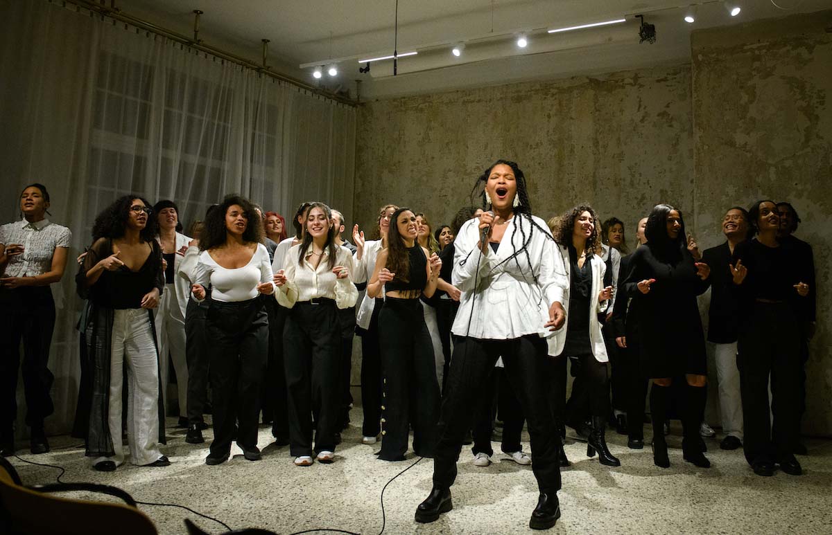 In the foreground, a female singer with long black dreadlocks, a white blouse and black trousers can be seen singing into the microphone in her hand with her mouth wide open. Behind her is a group of singers - at first glance most of them are identified as female - whose bodies are in motion, snapping their fingers or clapping their hands. The 25 or so members of the choir project A Song for You all wear black and white clothes in different styles. They stand in a room with an exposed concrete wall, a transparent white curtain at the window, a grey floor and are illuminated by small ceiling spotlights.