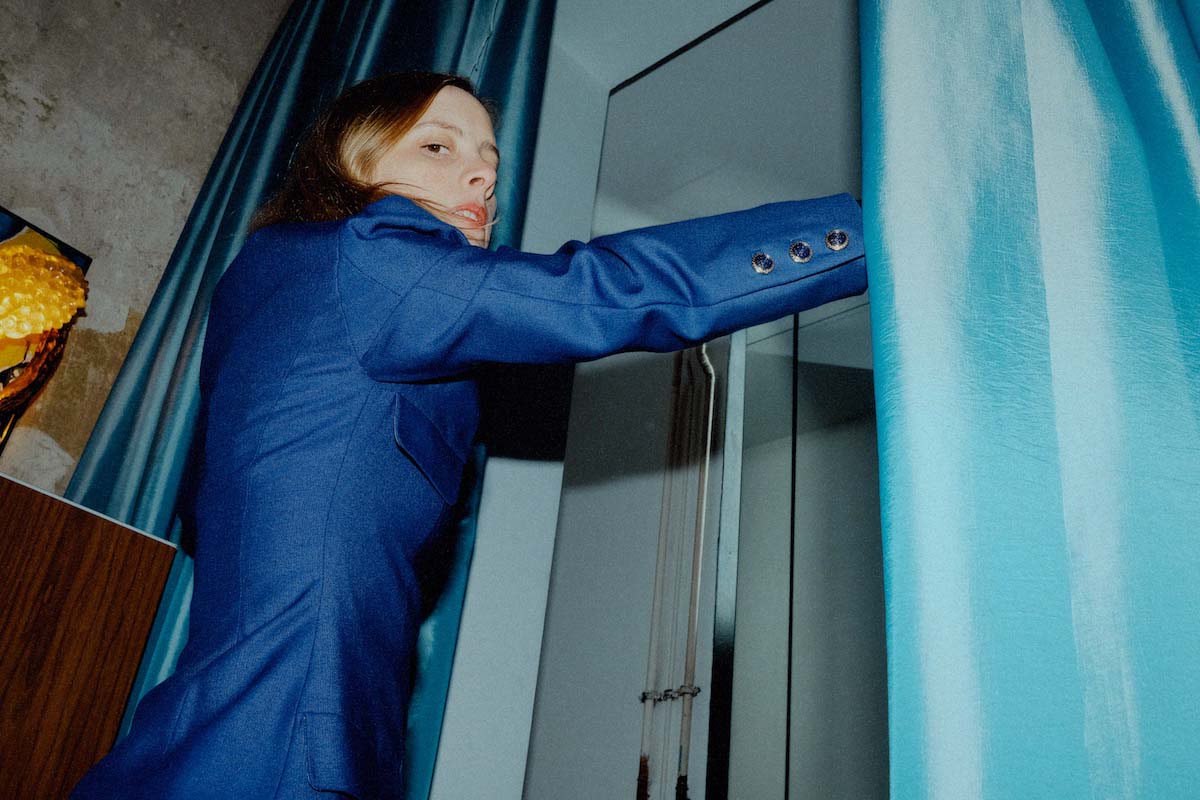 White, female person with shoulder-length brown hair is seen from diagonally behind pushing apart a curtain. Anika's visible right arm is outstretched, her hand hidden behind the light blue curtain. She wears a blue blazer and looks over her shoulder into the camera.