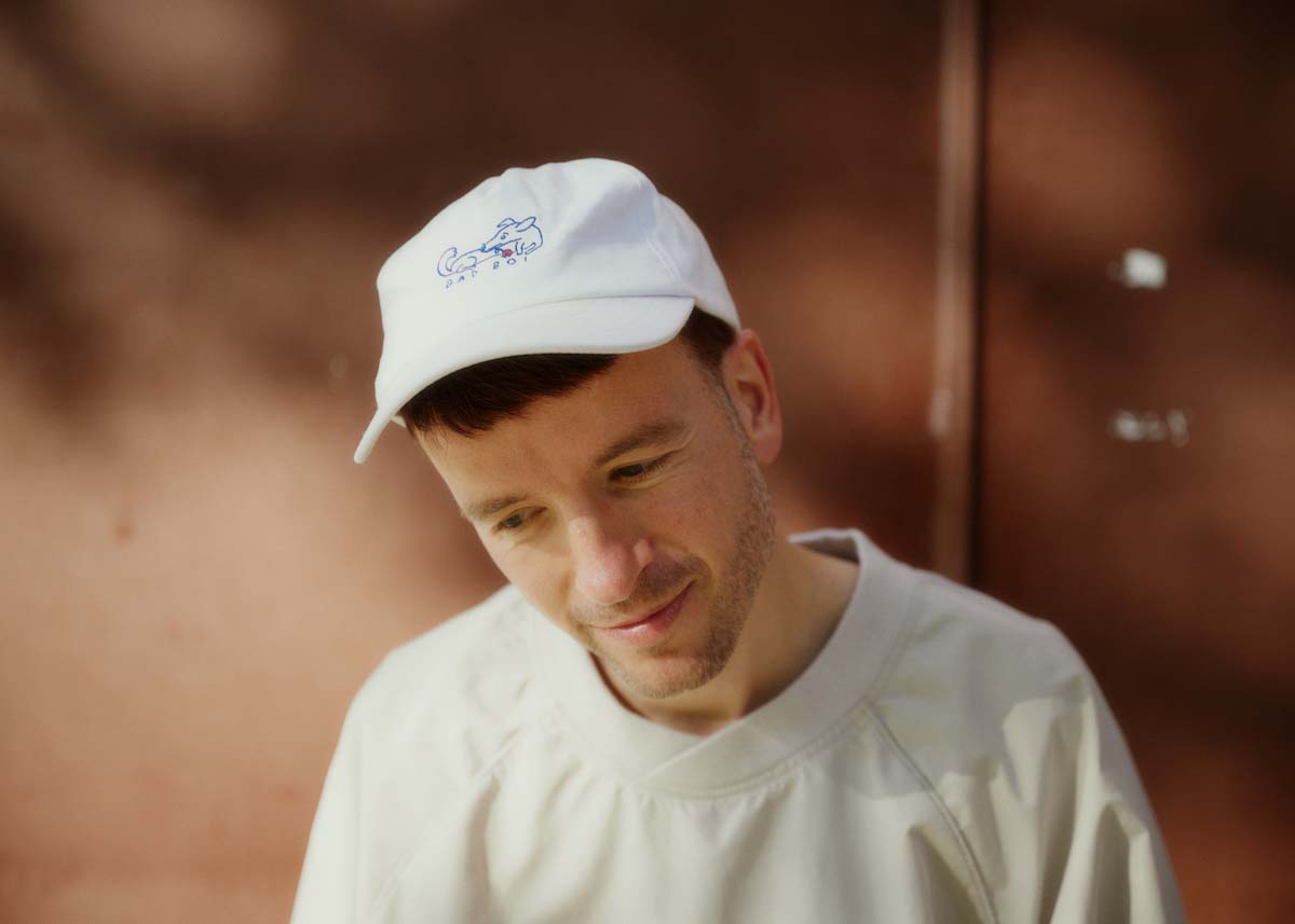 Shot of Robert Kretzschmar, in which he can be seen up to his chest. He is wearing a white baseball cap with short brown hair peeking out from underneath and a creamy white T-shirt. The white young man tilts his head slightly forward and looks out of the picture below. In the background, a brown wall can be seen out of focus.