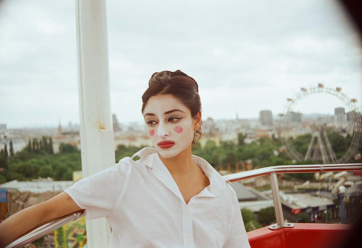 Young woman in a white blouse and brown pinned-up hair. Sophie Royer has her face painted completely white like a clown, with red dots on her cheekbones and red lips. She sits sadly looking to the side high up in the gondola of a ferris wheel, of which only a small part can be seen. In the background, too, a ferris wheel can be seen out of focus in the distance, as well as a view over a city.