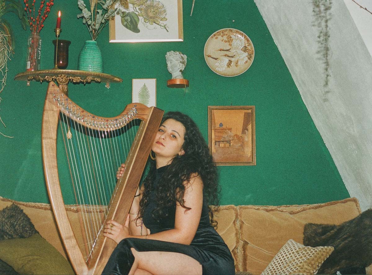 Young woman with long curly hair and a sleeveless, black, skin-tight dress whose high slit exposes her leg as she crosses her legs. Zainab Lax is sitting on a brown couch with various throw pillows on it, holding a harp in both hands with her face nestled against it. She looks into the camera. Behind her is a green wall with various decorative elements - pictures, a small bust, a decorative plate, a shelf with vases and a candle.