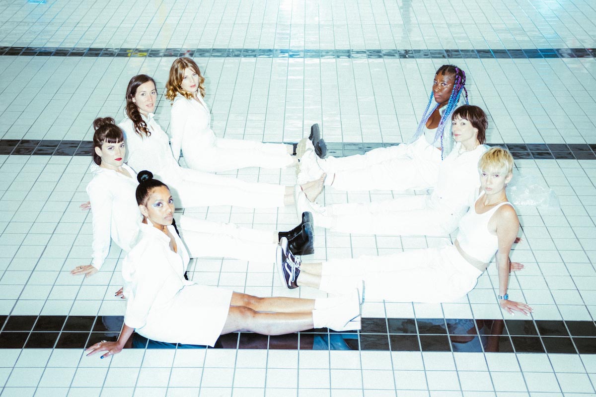 Seven diverse, young women, the FEM*ERGY COLLECTIVE, sit on a white tiled floor, which is permeated with parallel black tile stripes. Four sit on the left, opposite the other three on the right, all with their legs stretched out, all wearing white clothes in different styles, supporting themselves with their hands behind them and looking quite seriously at the camera.