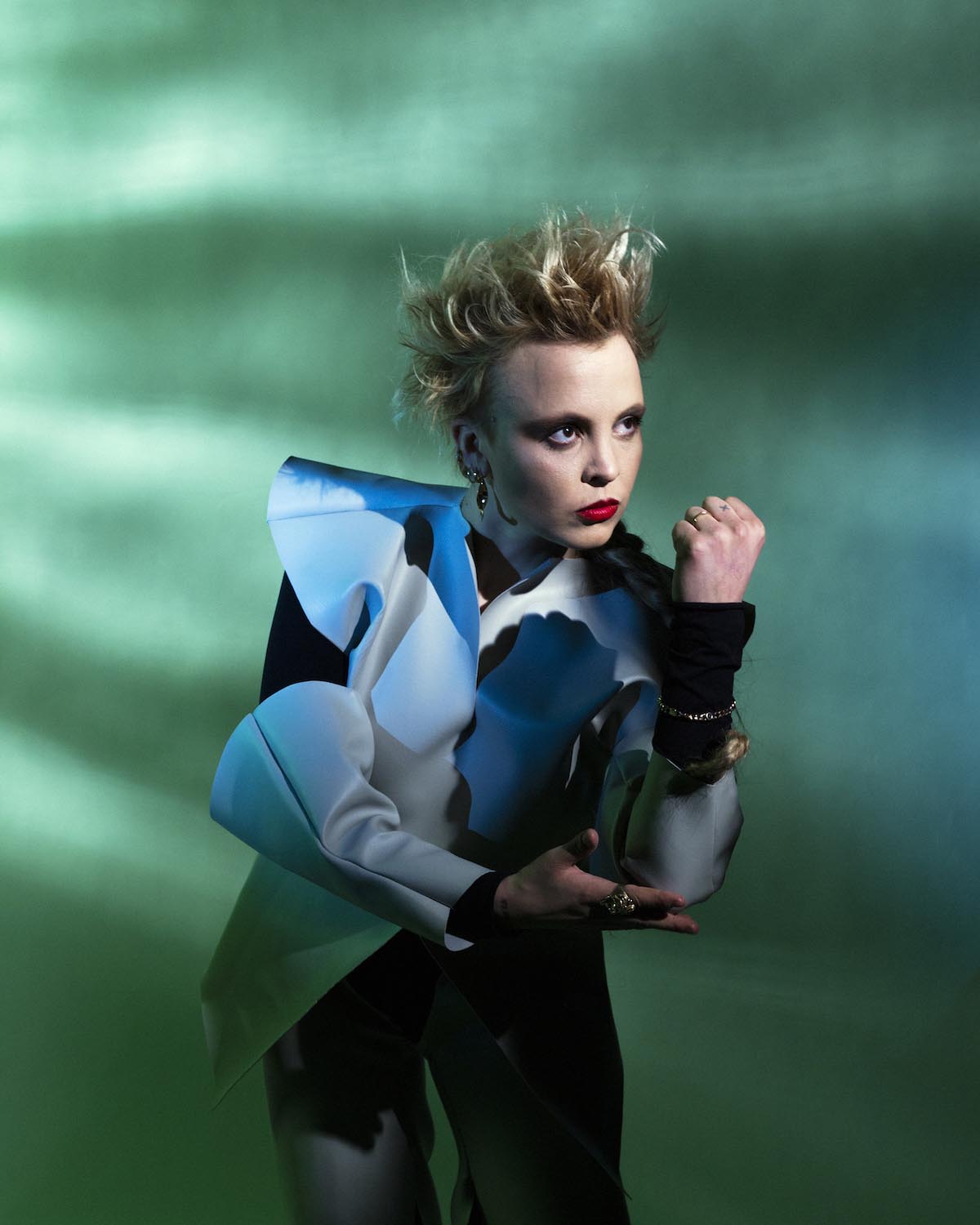 Young white woman with short blond hair styled upwards, dark eye make-up and red lips looks combatively out of the picture to the right. L Twills has her left hand clenched into a fist and holds her arm bent upwards, supporting her elbow with the other hand. She is wearing an unusual jacket with a black base and white wide set shoulders and white trumpet-shaped sleeve patches on the forearm. The background shimmers greenish with shadows that also fall on L Twills.