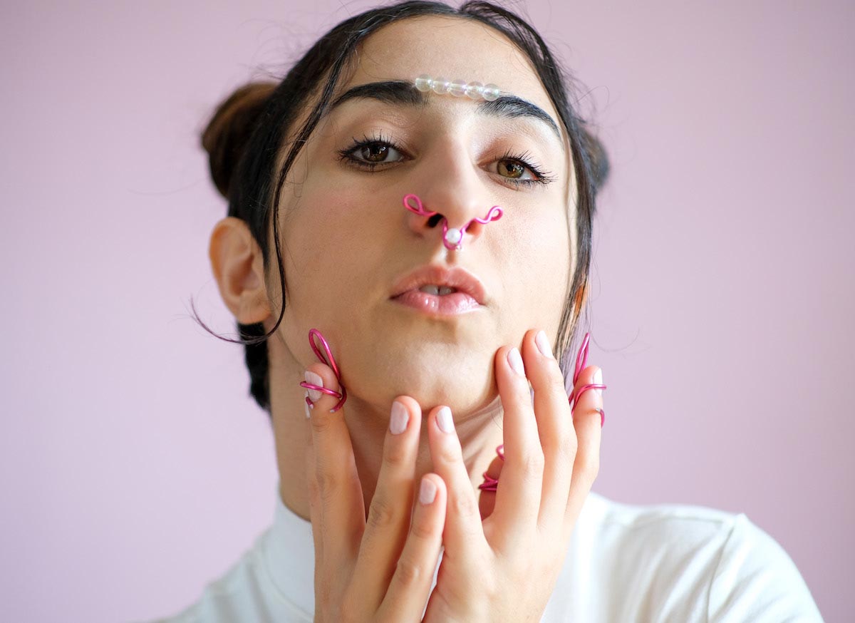 Porträtfoto einen jungen Frau mit schwarzen Haaren, die zu Space Buns gebunden sind. MADANII blickt direkt in die Kamera und hat die Lippen leicht geöffnet. Zwischen ihren schwarzen Augenbrauen sind fünf transparente Perlen in einer Reihe befestigt. Sie berührt ihr Kinn filigran mit beiden Händen. In den Nasenlöchern und an zwei Fingern hat sie sich pinkfarbene geschwungene Drahtgebilde befestigt.