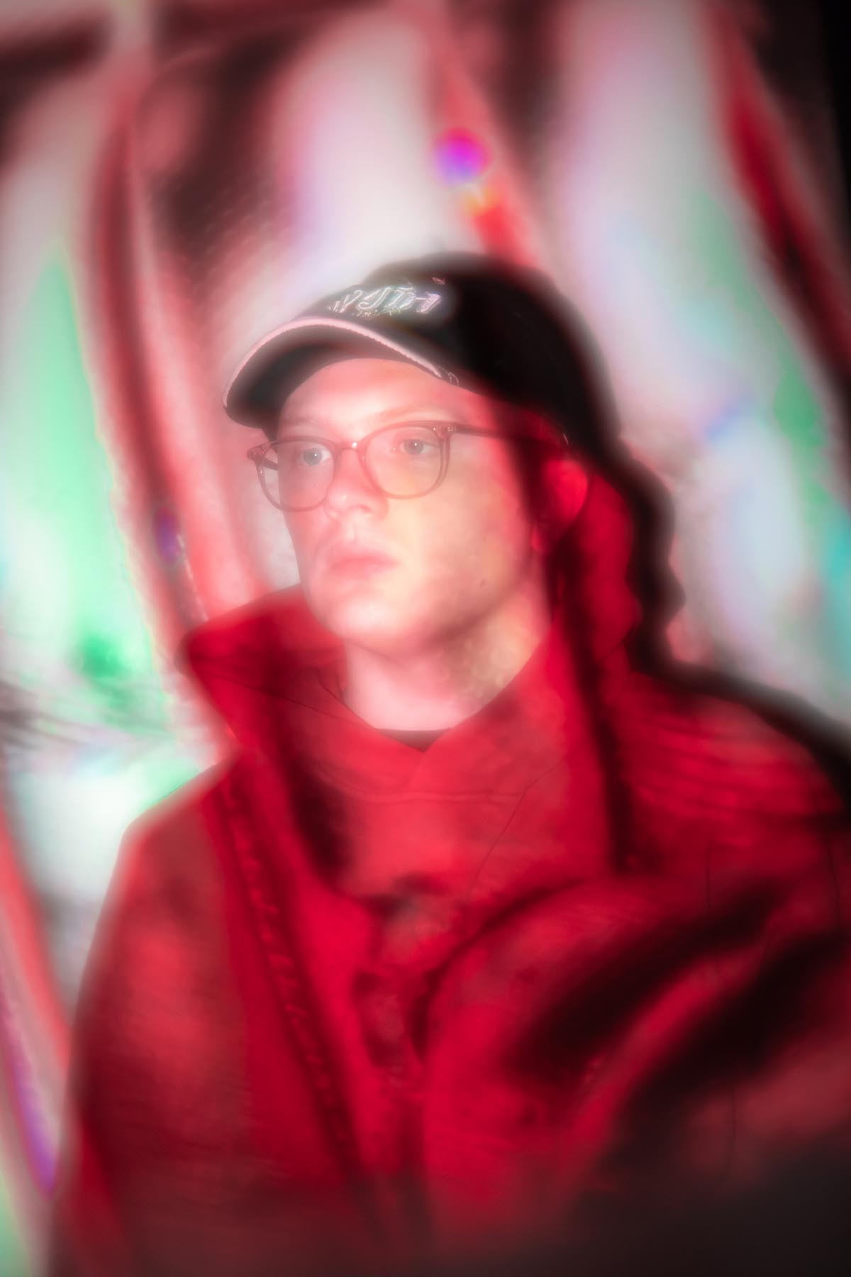 Young, white man with glasses and a black baseball cap with a white border on the shield wears a red hooded jumper and looks out of the picture very serious to the left. umru's torso can be seen, he is standing in front of a wall and something is projected onto him that cannot be seen and looks like a coloured veil on the picture.
